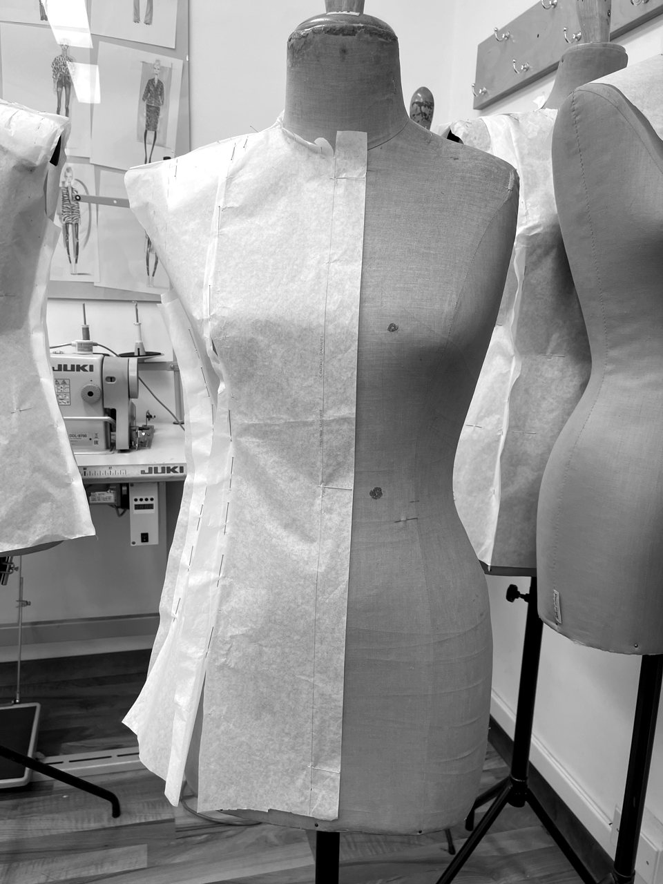 A bodice half-draped in pattern paper on the form, one side worked and one side bare, sewing machines and croquis behind