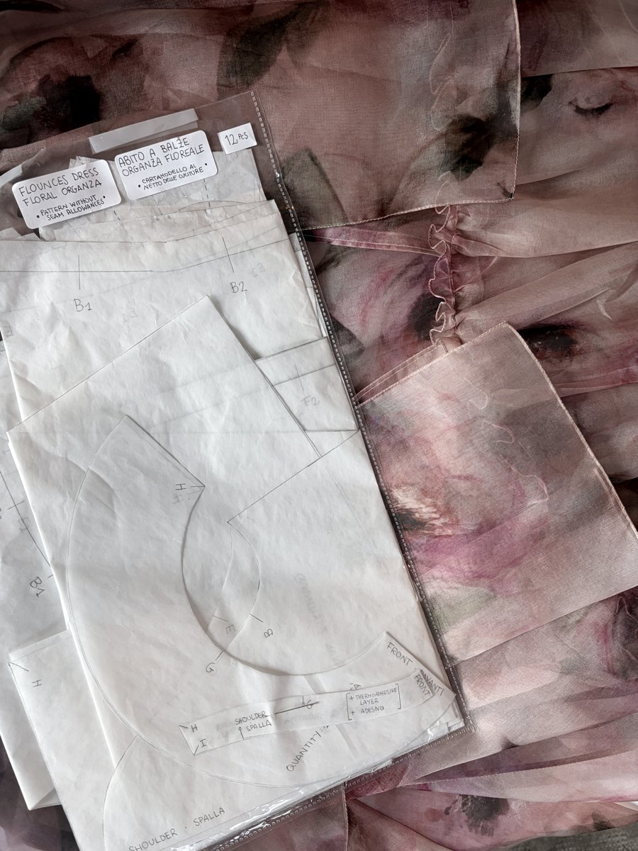 A folded couture pattern in its sleeve, labelled "flounces dress, floral organza, pattern without seam allowances" in English and Italian, resting on the finished floral silk dress it was drafted for
