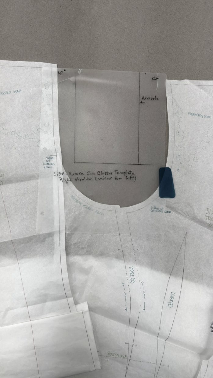 A hand-drafted couture pattern laid out on the cutting table: a bodice front with the centre front, armhole, neck point, darts and buttonhole marked by hand, seam allowances and balance notches along every seam, and handwritten notes for the embroidery zone and construction