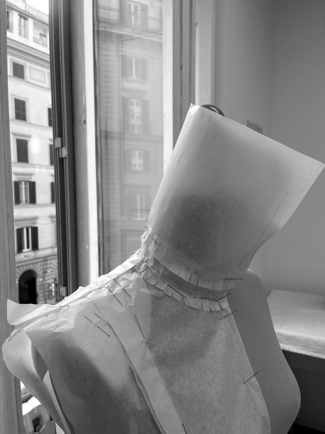 Pattern paper draped and pinned on a dress form in our Made in Italy atelier, a tall window behind it looking out onto a classical Roman building