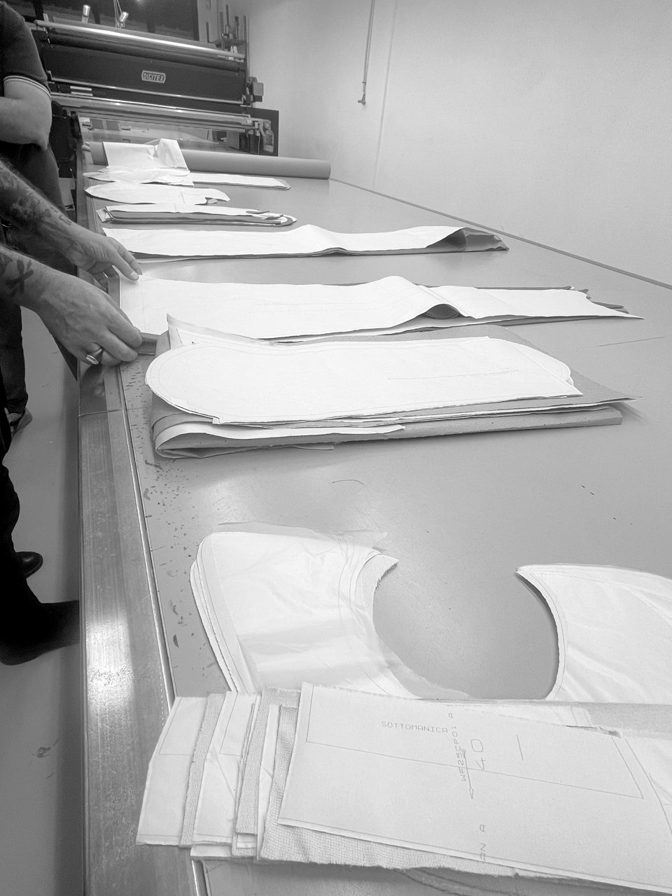 Several full paper patterns laid out along a long metal cutting table, stacked and in progress, a patternmaker working over them